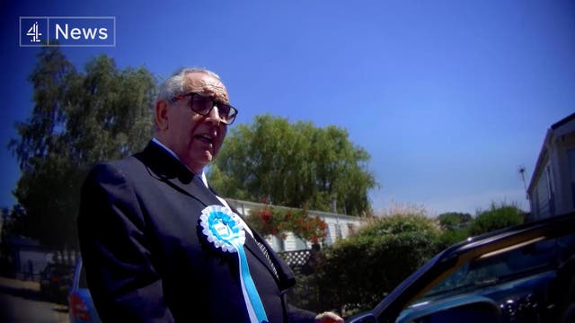 A man standing against a blue sky wearing a Reform UK rosette 
