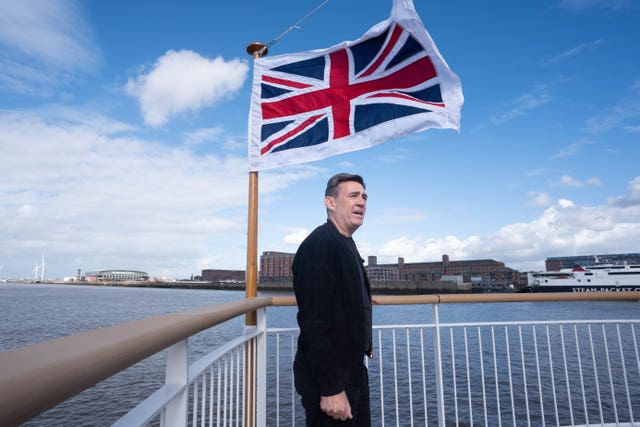 Andy Burnham pictured standing under a British flag that is flying in the wind, at a port