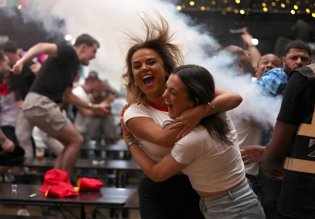 Two female England fans hug and a man jumps on a bench in the background as the crowd at BOXPark Wembley celebrate Jude Bellingham's equaliser against Slovakia