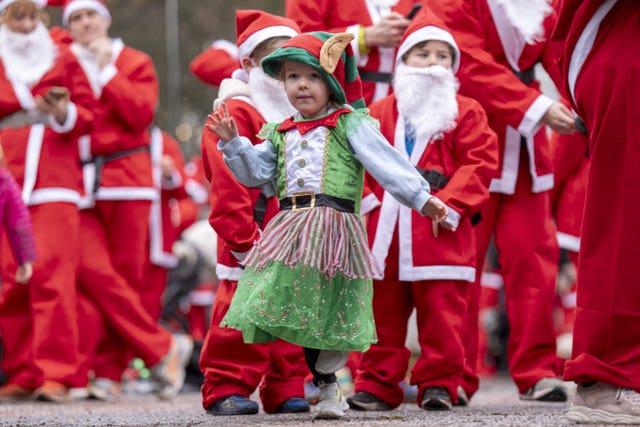 A young child in an elf outfit dances in front of Santas