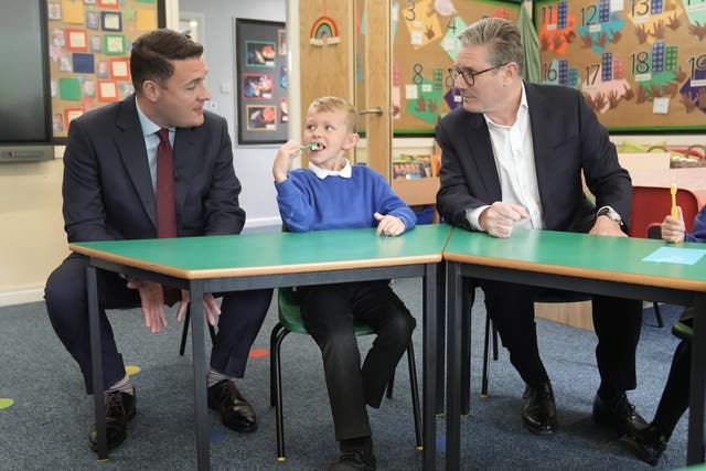 Shadow health secretary Wes Streeting and Sir Keir Starmer sit either side of a young blond boy in school uniform sitting at a low desk in a classroom 