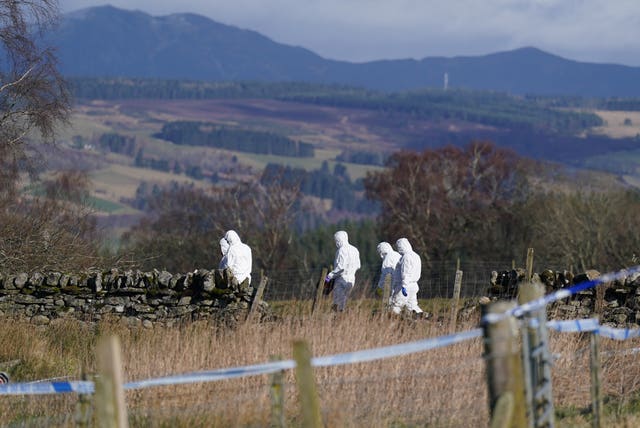 Police in white forensic suits walk in a rural area, with police tape cordoning the area off in the foreground