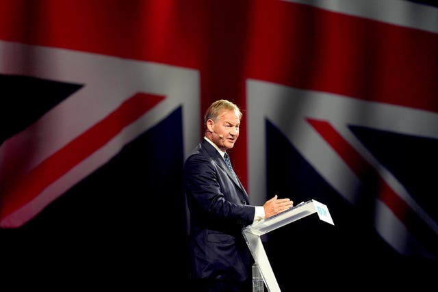 Rupert Lowe making a speech from a podium in front of a giant Union flag