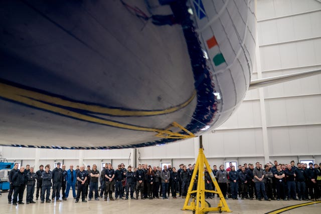 A large group of people in an aircraft hangar, with the nose of a plane above