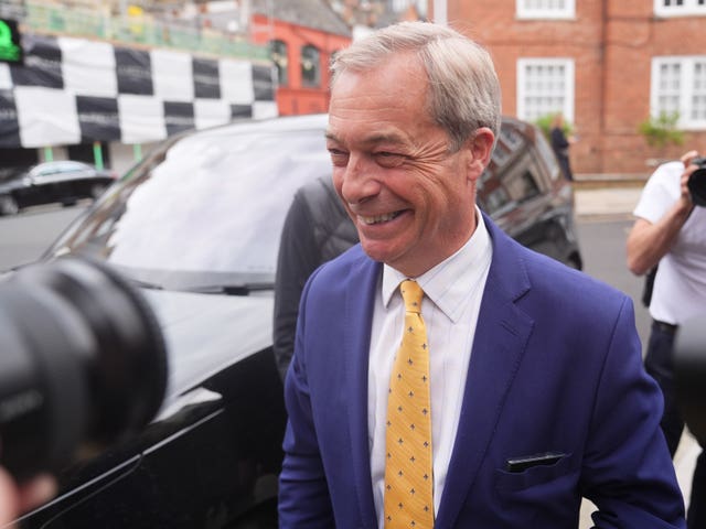 Nigel Farage smiling, standing outside near a car while surrounded by photographers