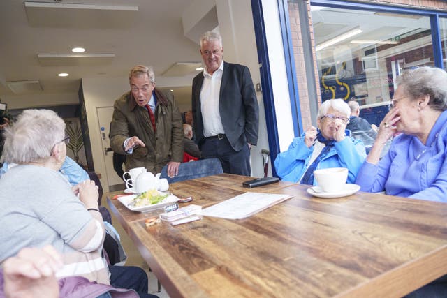 Nigel Farage leans down to talk to an elderly lady over a table holding a salad and tea pot while Lee Anderson watches with his hands in his pockets, and two women dressed in blue laugh 