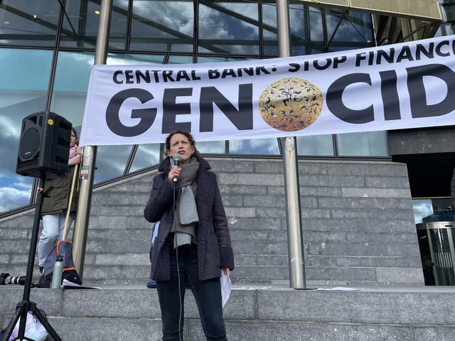 Marie Sherlock speaking into a microphone, below a banner reading 'Central Bank: Stop financing genocide'