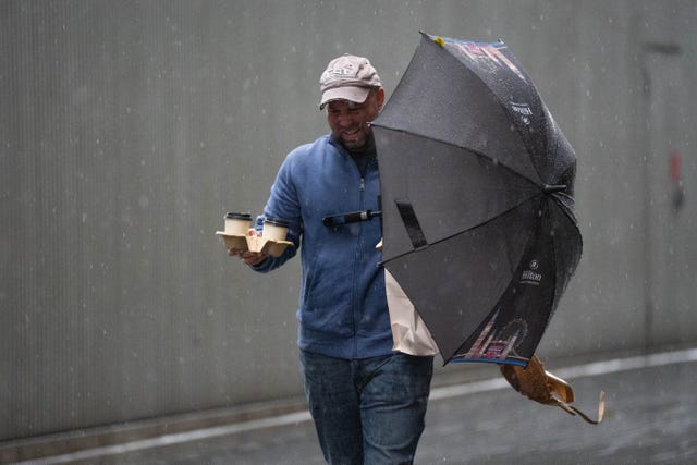 A man struggles to carry some takeaway coffee and an umbrella near Tower Bridge in London