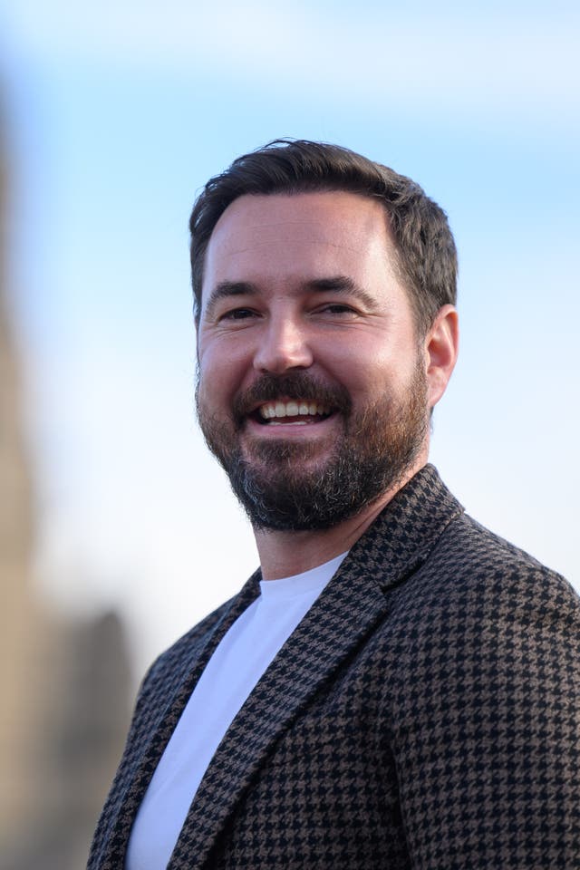 Martin Compston wearing a houndstooth blazer and white T-shirt while standing outside, smiling at the camera