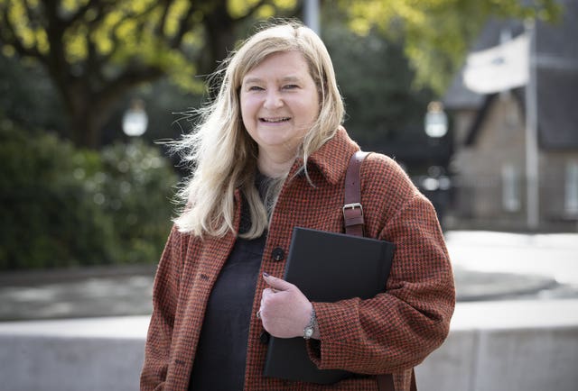 Public Health Secretary Jenni Minto smiling at the camera while walking in a leafy street on a sunny day. She is wearing a red coat and has a folder under her arm.