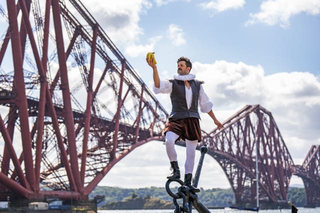 Performer stands on anchor, looking at bananas in his hand, with the Forth Bridge in the background