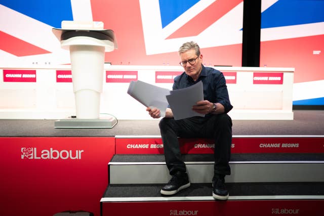 Sir Keir Starmer reads papers while sitting on stage emblazoned with Labour signage