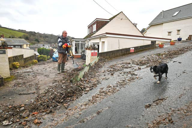 A dog walks past Paul Davis as he shovels debris from his driveway on Corner Lane, Combe Martin, North Devon, after flooding affected parts of the South West