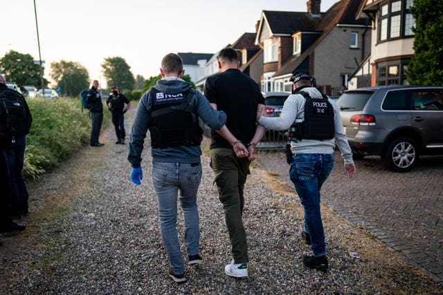 The back of a handcuffed man walking between two NCA officers, again wearing black vests with NCA written on the back.