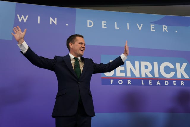 Robert Jenrick waves on stage at his leadership campaign launch. He stands in front of a large banner reading "Win. Deliver. Jenrick for Leader"