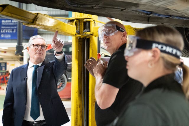 Sir Keir Starmer points to the underside of a car watched by a man and and a woman in protective goggles