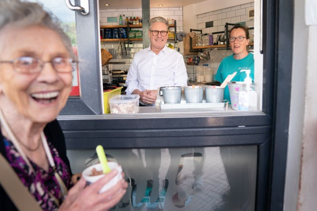 An older woman smiles in the foreground while holding an ice cream while Sir Keir Starmer smiles in the background inside an ice cream parlour