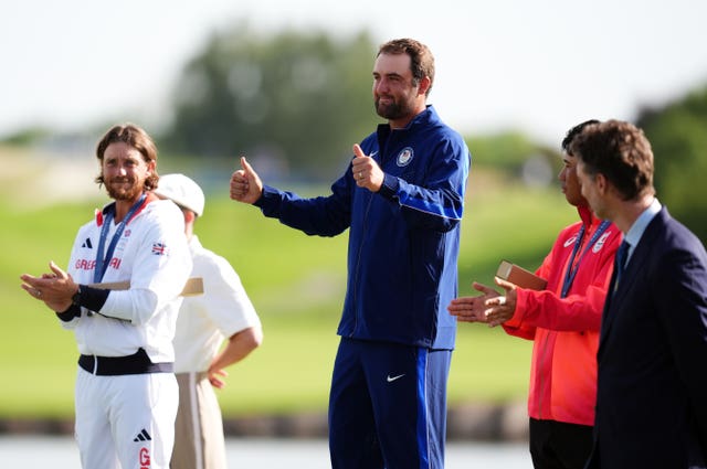 USA’s Scottie Scheffler gives the thumbs up on the podium after winning a gold medal, with Tommy Fleetwood on his right and Hideki Matsuyama on his left. 