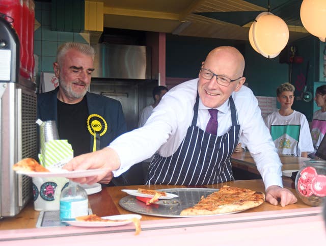 John Swinney in a blue and white striped apron passes a plate with a slice of pizza from behind a serving counter
