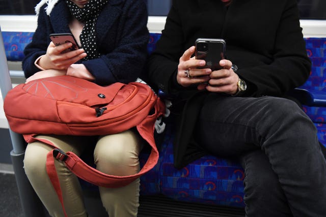 A close-up of an unidentifiable man and woman sitting next to each other on the Tube, both looking at their phones.