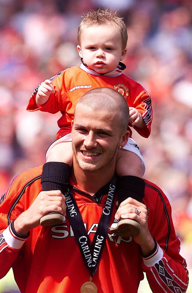 David Beckham wearing a Manchester United shirt and with a Premiership medal around his neck holding a young Brooklyn Beckham on his shoulders