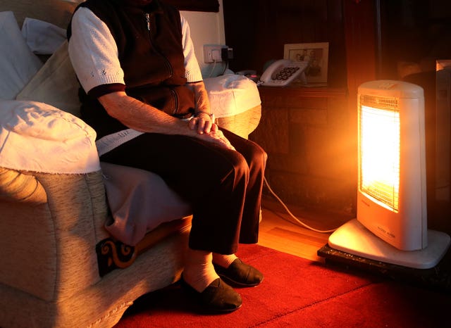 An elderly woman sits next to an electric heater 