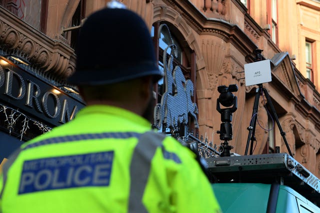 The back of a uniformed officer, looking up at a facial recognition camera that is mounted on the roof of a green van. 