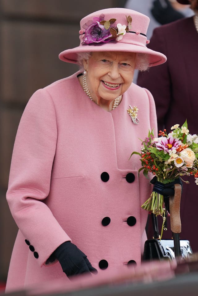Queen Elizabeth II smiling while wearing a pink coat and matching hat, and carrying a posy of flowers