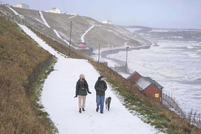 Two people wrapped in warm clothing walking a dog in snowy conditions on Whitby seafront in Yorkshire