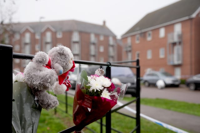 Flowers and a teddy bear attached to a fence in front of blocks of flats