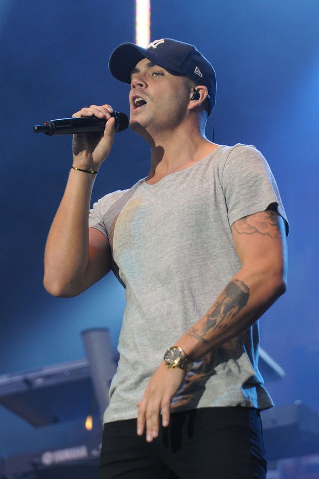 Max George performing on stage, wearing a grey T-shirt, black trousers and a blue baseball cap