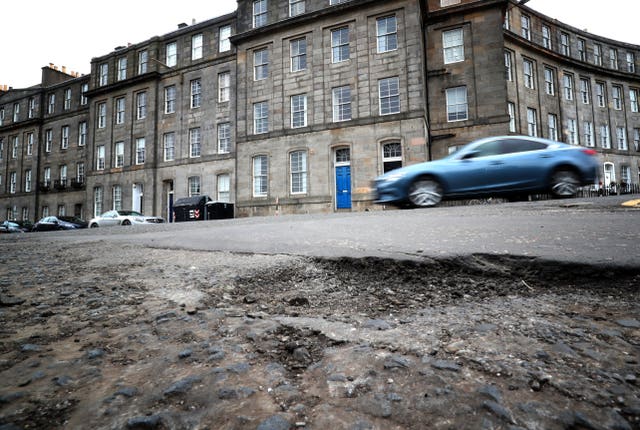 A blue car drives past a big pothole in a road