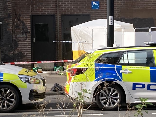 A white forensics tent behind police tape, with part of two parked police cars in front.