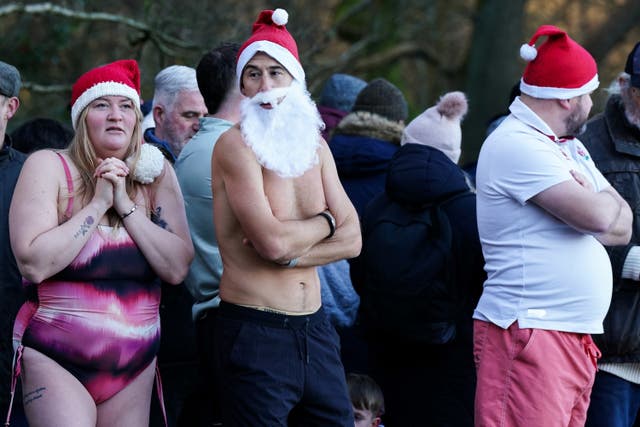 A topless man wearing a Santa beard and a Christmas hat folds his arms while standing next to a woman in a swimsuit and Christmas hat looking cold