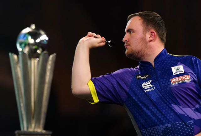 Luke Littler throws alongside the World Championship trophy