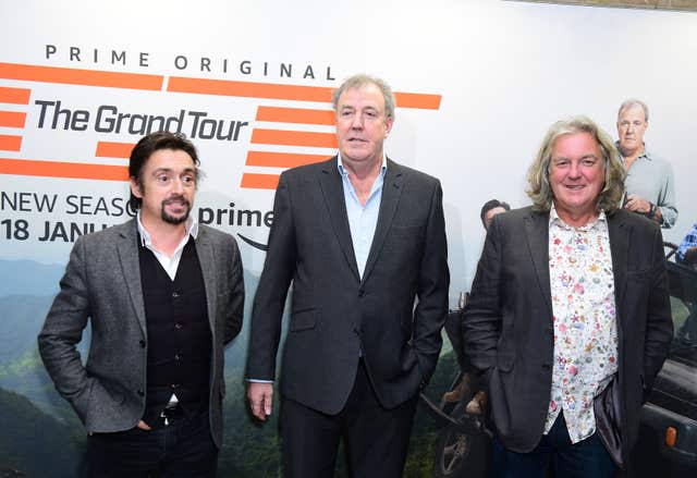 Richard Hammond, Jeremy Clarkson and James May  standing in suits in front of a Grand Tour sign