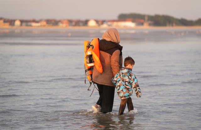 A wide shot of the sea, a woman holding a lifejacket over her shoulder holds the hand of a young boy. They are wading through the water up to their knees