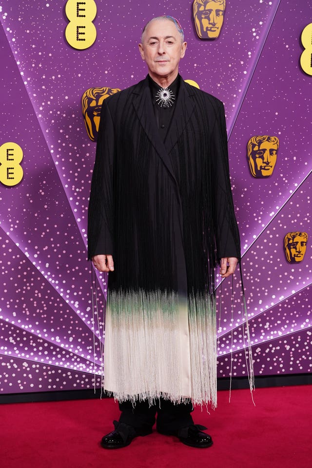 Alan Cumming in a long fringed coat with ombre effect ranging from black at his shoulders to white at the hem