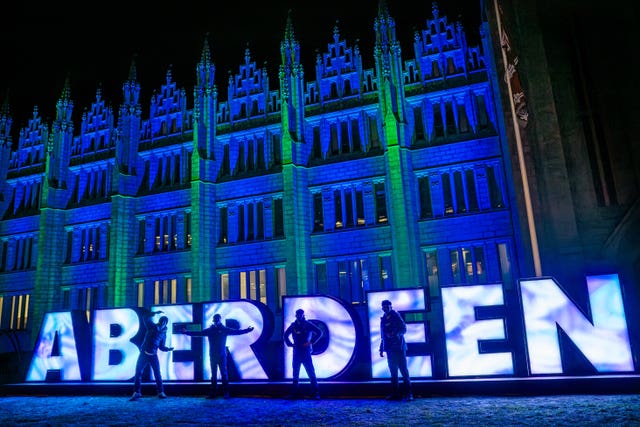 Four people standing in front of giant letters that spell Aberdeen with a building lit in blue behind her