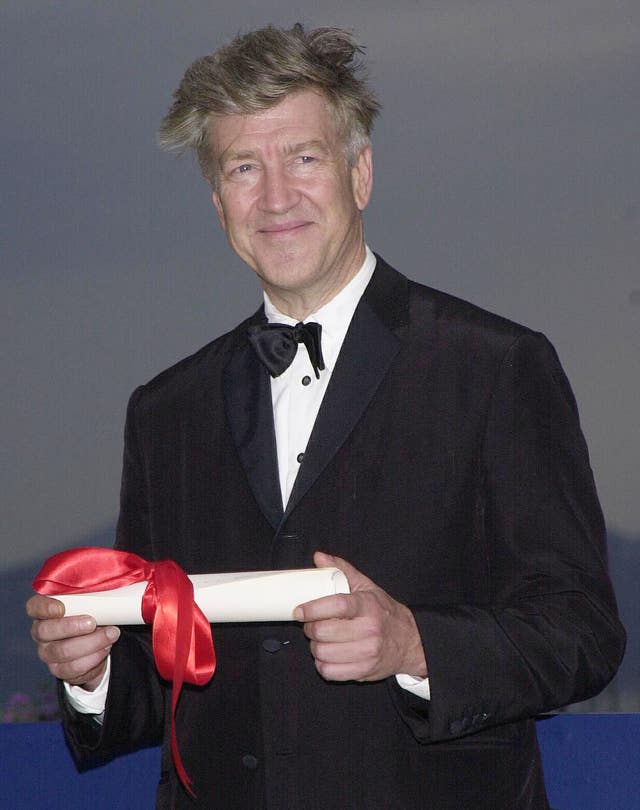 David Lynch, wearing a tuxedo, smiles while holding a scroll of paper wrapped in a red ribbon