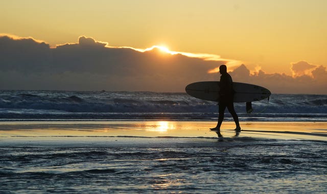 A surfer carries their board along the beach in front of a low sun