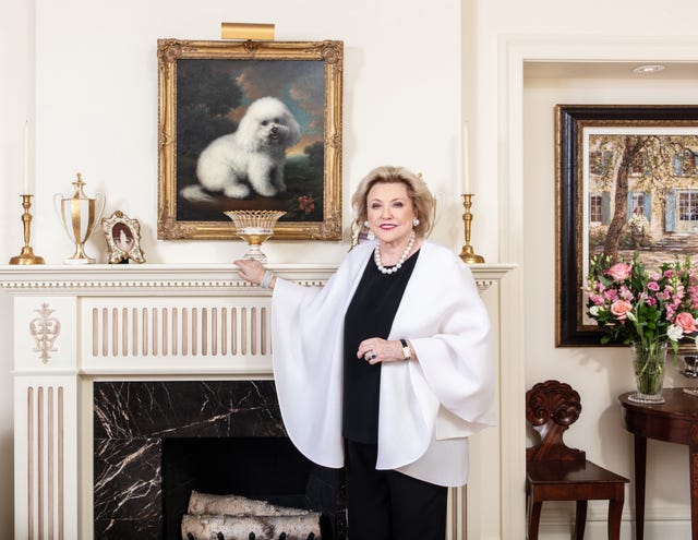 Barbara Taylor Bradford in a white wrap standing in front of a fireplace