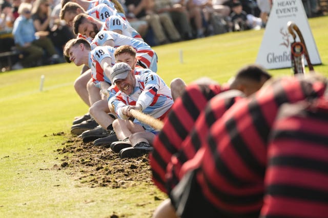 A team of men wearing black and red-striped tops in the foreground pulls on a rope while a team dressed in blue, white and red pulls on the same rope in the background as they compete in a tug of war