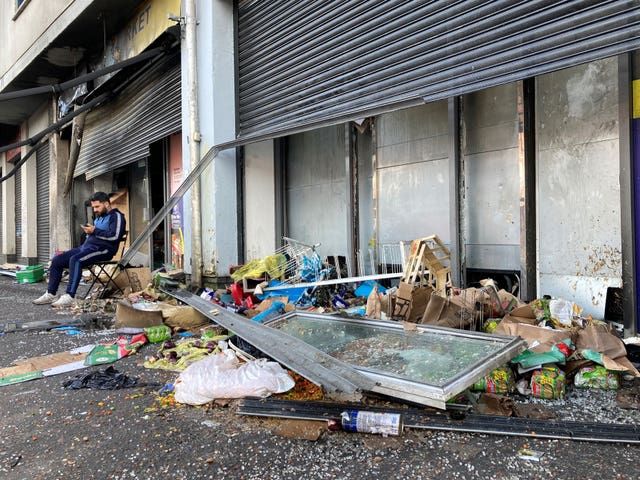 A man sits on a chair outside a damaged shop