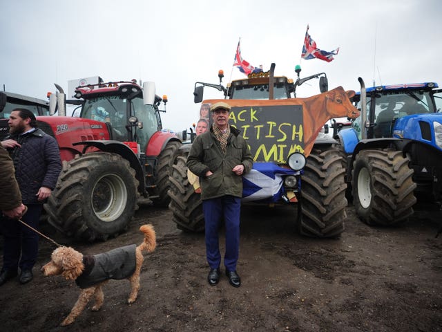 Nigel Farage wearing a coat, flat cap and scarf while standing in front of tractors