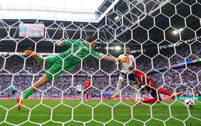 Photo from behind the goal, shows the netting and beyonbd that, a goalkeeper in green stretches to save a shot. The ball is in the bottom right and two players, one in white and one is red, stretch to hit the ball