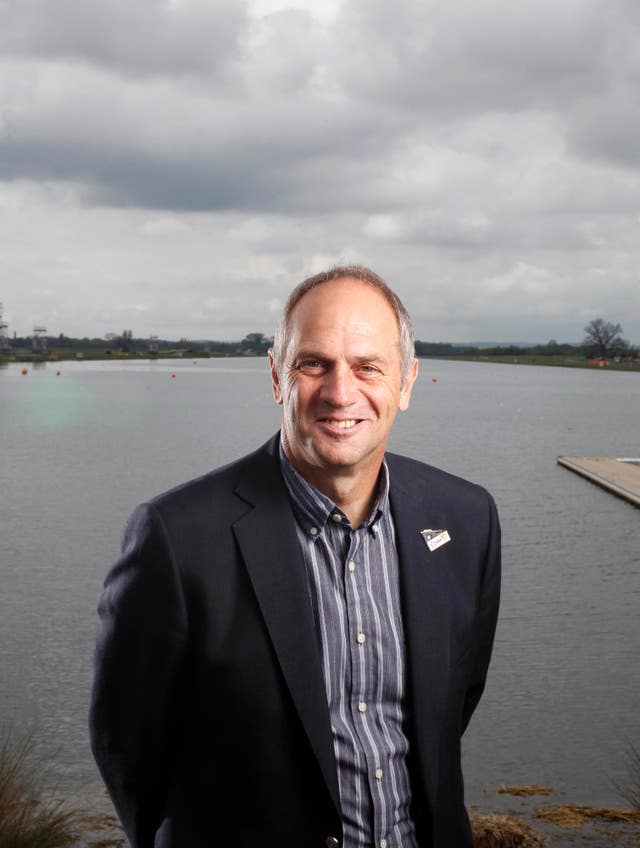 Sir Steve Redgrave stands in front of a river wearing a shirt and suit jacket, smiling at the camera
