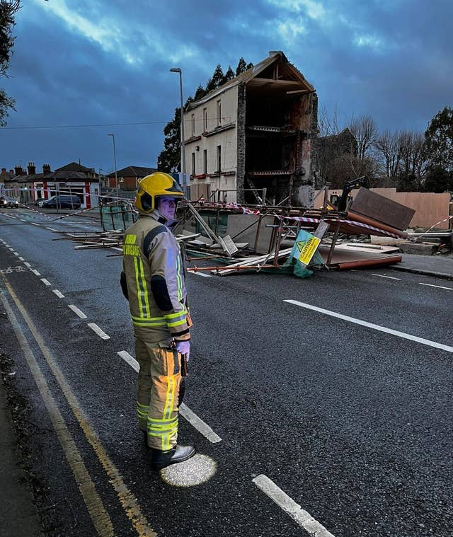 A firefighter in hi-vis stands in the middle of the road. Behind him a wall of scaffolding has collapsed into the road