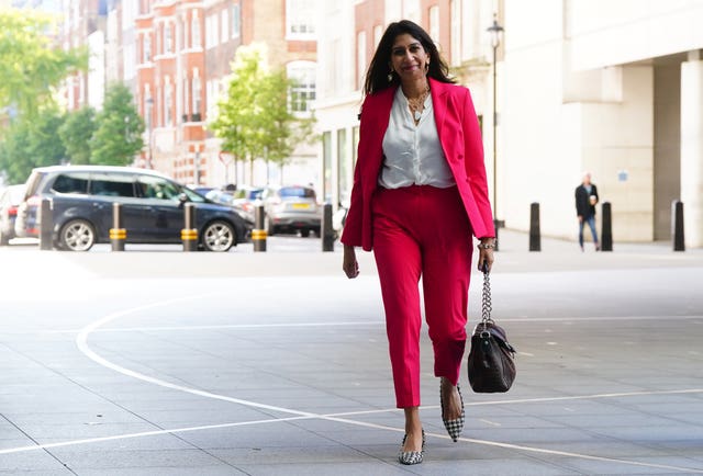 Suella Braverman wearing a red trouser suit, smiling as she walks outside