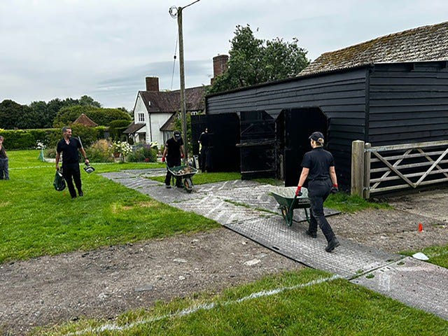 Two men wheel wheelbarrows near a barn